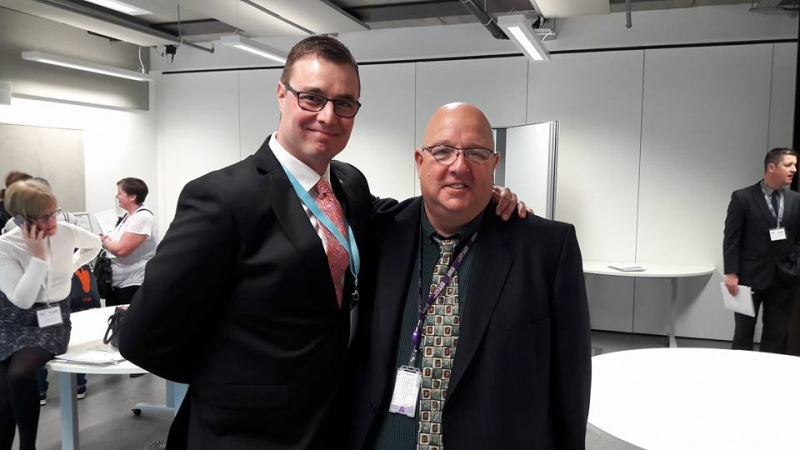 Kuva: Mikko Siljander / Mikko Siljander ja Mark Pollitt Glasgow'ssa Kaksi henkilöä puvuissa poseeraa kameralle vierekkäin. Taustalla on muita henkilöitä. Joku puhuu puhelimessa.